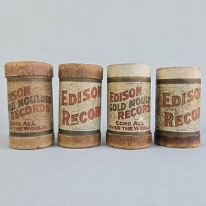 May include: Four cylindrical Edison Gold Moulded Records containers made of aged, light brown cardboard. Each container features a label with the text Edison Gold Moulded Records and Echo All Over The World in red and black ink with decorative flourishes.