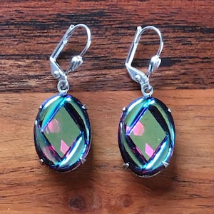 May include: A pair of iridescent oval earrings with silver-colored lever-back closures. The earrings feature a central diamond-shaped design within the oval, showcasing a blend of green, blue, and pink hues. The earrings are displayed on a wooden surface.