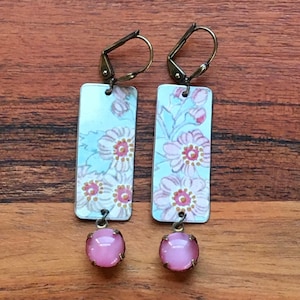 May include: A pair of dangle earrings with a light blue background and pink floral pattern. The earrings have a pink glass bead at the bottom.