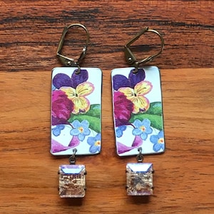 May include: A pair of rectangular earrings with a floral design. The earrings feature a white background with purple, yellow, and blue flowers. Each earring has a clear, rectangular crystal at the bottom and gold-colored ear wires.