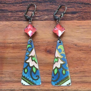 May include: A pair of dangle earrings with a blue, green, and pink patterned triangle pendant and a pink crystal at the top. The earrings have a silver-toned clip-on closure.