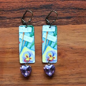 May include: A pair of rectangular earrings with a botanical theme. Each earring features a watercolor design with a pansy, the words "Sage" and "Thyme", and a purple heart-shaped charm. The earrings have bronze-colored ear wires.