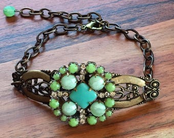 May include: A green and turquoise statement bracelet with a large, ornate, floral-shaped centerpiece. The bracelet is made of metal and has a chain link closure.