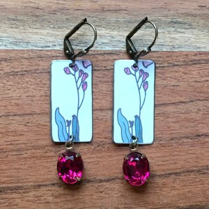 May include: A pair of dangle earrings with a white rectangular charm featuring a pink floral design and a dangling pink oval-shaped rhinestone.
