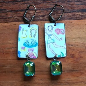 May include: A pair of earrings with a rectangular charm featuring a paper doll design and a green gemstone dangle. The paper doll charm has a white background with colorful details and a drawing of a girl in a dress.