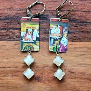 May include: A pair of earrings with a rectangular image of a bakery scene and two white, diamond-shaped beads hanging from each. The earrings have gold-toned hooks.