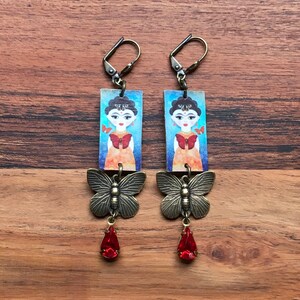 May include: A pair of dangle earrings with a rectangular charm featuring a colorful illustration of a woman with butterflies. The charm is attached to a bronze butterfly charm and a red teardrop crystal.