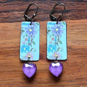 May include: A pair of dangle earrings. Each earring features a rectangular, light blue pendant with a floral design in purple, blue, and pink. A heart-shaped, purple gem hangs below each pendant. The earrings have silver-colored lever-back closures.