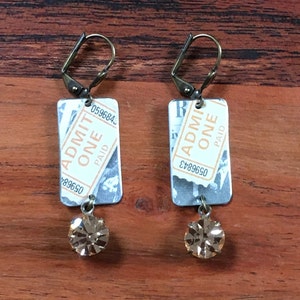 May include: A pair of earrings featuring vintage-style ticket charms with the text "ADMIT ONE PAID" and a small, clear crystal dangle. The charms are attached to gold-toned ear wires.