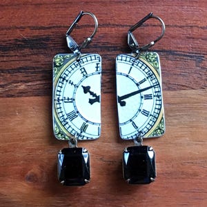 May include: A pair of rectangular earrings featuring a clock face design with black hands and Roman numerals. The earrings have a black square gemstone at the bottom and silver-tone ear wires. The clock face has a cream background with a gold border.