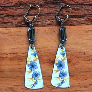 May include: A pair of dangle earrings with a white background and blue floral pattern. The earrings feature a dark blue crystal bead and a silver hook.