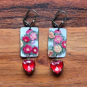 May include: A pair of dangle earrings. Each earring features a rectangular floral design with red and pink flowers, topped with a red heart-shaped jewel. The earrings have a silver-colored lever back closure.