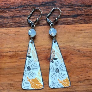 May include: A pair of dangle earrings with a floral design. The earrings feature a white background with a large white flower and orange and brown leaves. The earrings are triangular in shape and have a silver-colored hook.