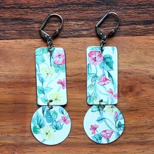 May include: A pair of floral earrings with a rectangular top and a circular bottom. The earrings feature a white background with pink, yellow, and green floral patterns. The earrings are connected with a small metal ring and have a silver lever-back closure.