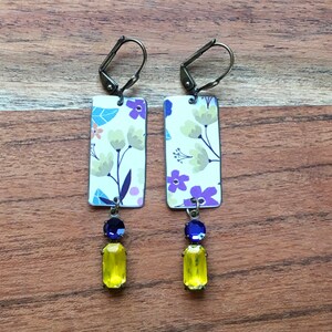 May include: A pair of dangle earrings with a floral pattern on a white background. The earrings have a yellow and blue gemstone dangle.