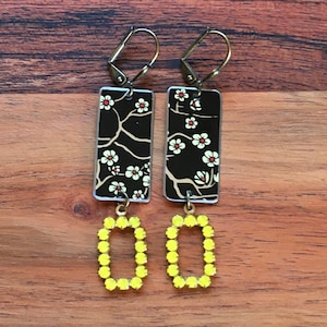 May include: A pair of dangle earrings with a black and white floral pattern on the top portion and a yellow rhinestone rectangle on the bottom portion.