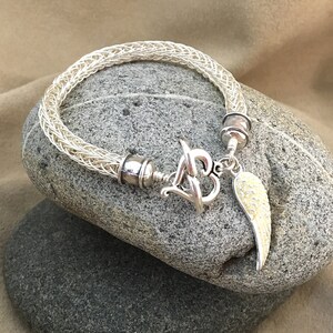 Op de afbeelding: Een witte gevlochten armband met een zilveren hartbedel en een zilveren engelenvleugelbedel. De armband ligt op een grijze steen.