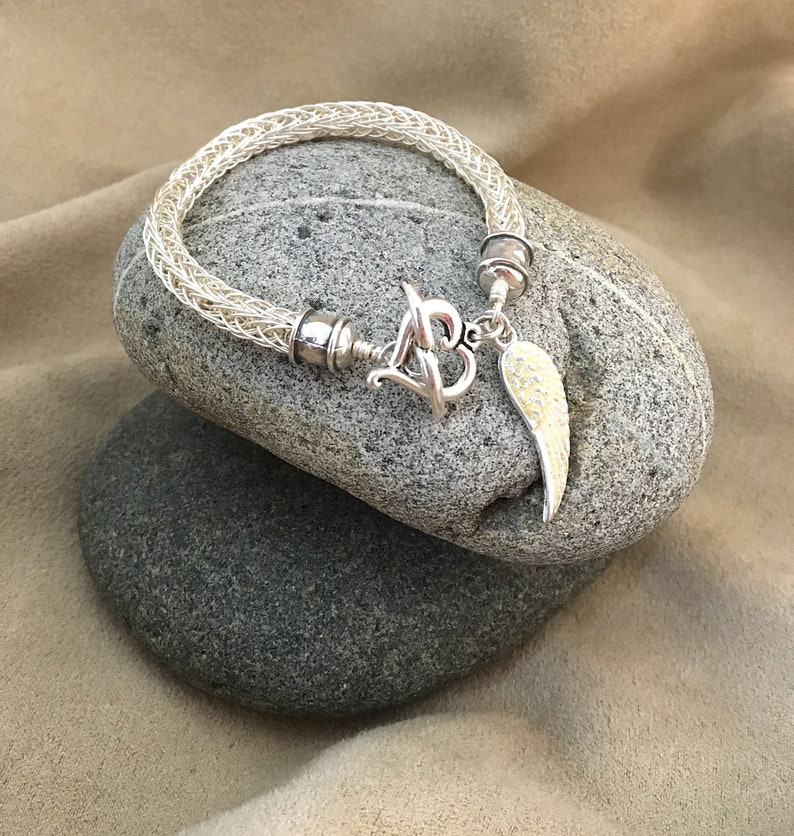 Op de afbeelding: Een witte gevlochten armband met een zilveren hartbedel en een zilveren engelenvleugelbedel. De armband ligt op een grijze steen.