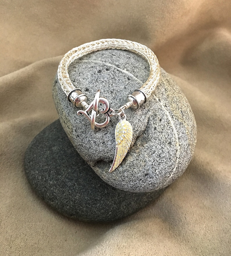 Op de afbeelding: Een zilveren armband met een wit gevlochten koord en een zilveren bedel met een enkele engelenvleugel. De bedel is bevestigd aan de armband met een zilveren sluiting.