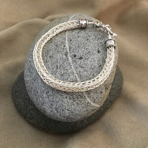 Op de afbeelding: Een zilveren draad armband met een geweven ontwerp en zilveren sluitingen. De armband wordt getoond op twee grijze stenen.