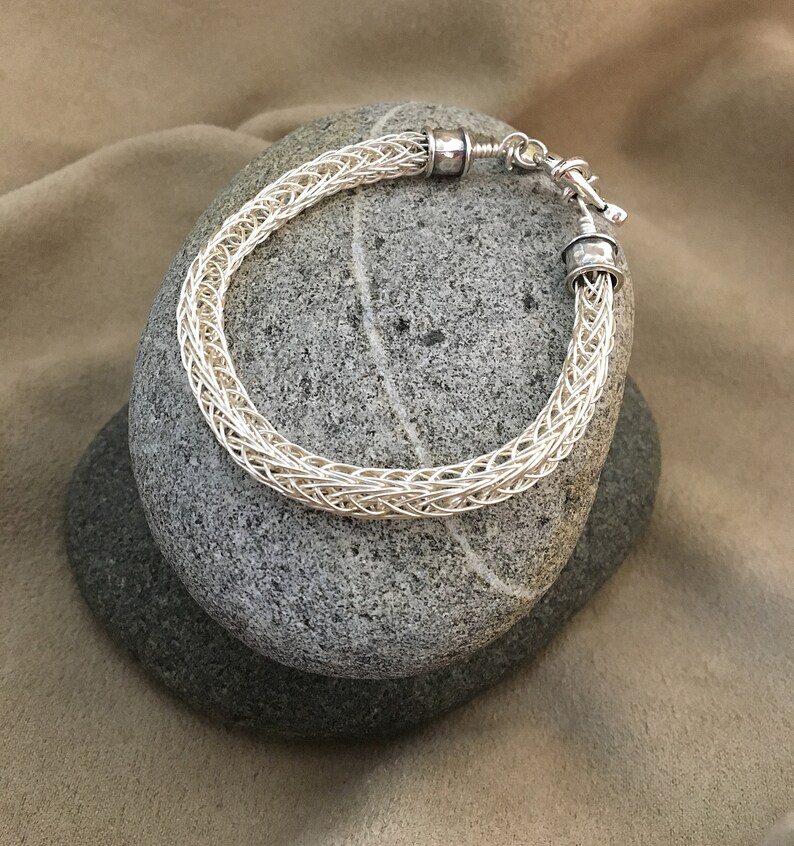 Op de afbeelding: Een zilveren draad armband met een geweven ontwerp en zilveren sluitingen. De armband wordt getoond op twee grijze stenen.