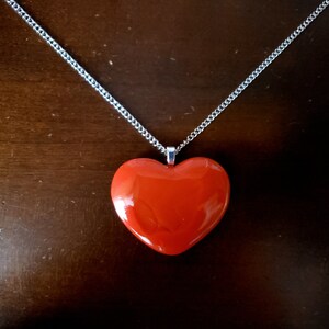 Puede incluir: Un collar con colgante en forma de coraz&oacute;n rojo y cadena plateada. El coraz&oacute;n es rojo liso y brillante, y la cadena es de color plata delicado. El collar se muestra sobre una superficie marr&oacute;n oscuro.