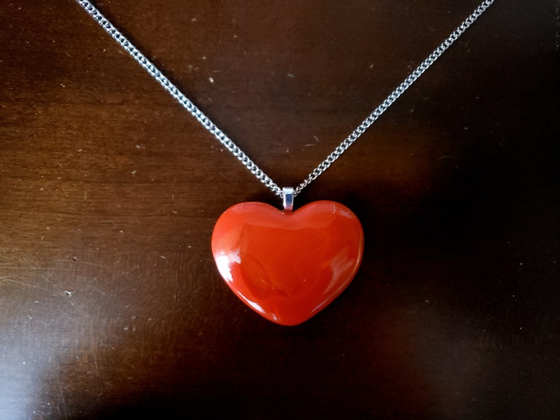 Puede incluir: Un collar con colgante en forma de coraz&oacute;n rojo y cadena plateada. El coraz&oacute;n es rojo liso y brillante, y la cadena es de color plata delicado. El collar se muestra sobre una superficie marr&oacute;n oscuro.