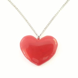 Puede incluir: Un collar con colgante en forma de coraz&oacute;n rojo y cadena plateada. El coraz&oacute;n es de color rojo brillante y liso, centrado sobre un fondo blanco. La cadena es una fina cadena de eslabones plateada.