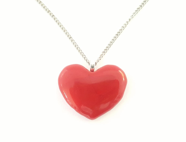 Puede incluir: Un collar con colgante en forma de coraz&oacute;n rojo y cadena plateada. El coraz&oacute;n es de color rojo brillante y liso, centrado sobre un fondo blanco. La cadena es una fina cadena de eslabones plateada.