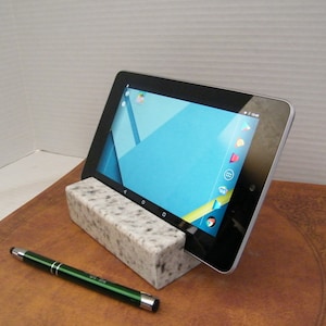 May include: A white granite tablet stand with a black tablet resting on top. The tablet is displaying the Android home screen. A green stylus pen is lying on a brown surface.