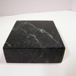 May include: A rectangular block made of dark black stone with prominent white veining throughout the surface. The object features a smooth, polished finish and sharp, clean edges, presenting a solid and uniform appearance from this perspective.