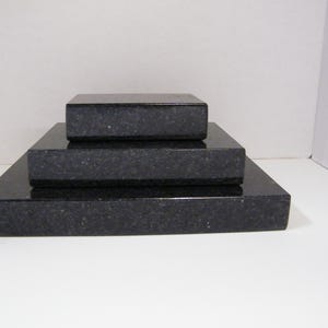 May include: Three stacked, rectangular black granite blocks. The blocks are of varying sizes, with the largest at the base and the smallest on top. The granite has a speckled texture and a polished finish.