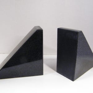Puede incluir: Dos sujetalibros de granito negro. Cada sujetalibros tiene forma de tri&aacute;ngulo rect&aacute;ngulo con una parte superior plana y un acabado pulido. Los sujetalibros se muestran sobre una superficie blanca.