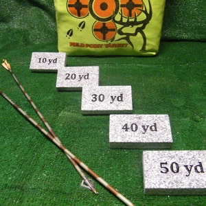 May include: Five gray rectangular blocks with black text indicating distances in yards: 10 yd, 20 yd, 30 yd, 40 yd, and 50 yd. Two camouflage arrows are lying on a green surface.