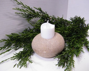 May include: A natural stone candle holder with a white candle. The candle holder is a rounded, beige stone with a hole to hold the candle. Green evergreen branches are arranged around the candle holder.