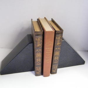 Puede incluir: Tres libros antiguos sostenidos entre dos sujetalibros triangulares negros. Los libros tienen cubiertas marrones y beige, con detalles decorativos dorados. El libro del medio es de color naranja claro.