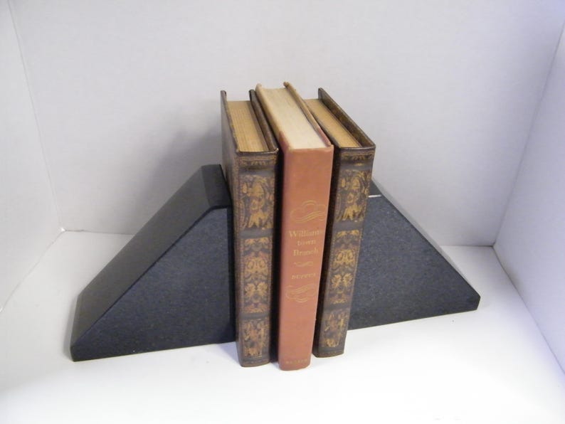 Puede incluir: Tres libros antiguos sostenidos entre dos sujetalibros triangulares negros. Los libros tienen cubiertas marrones y beige, con detalles decorativos dorados. El libro del medio es de color naranja claro.