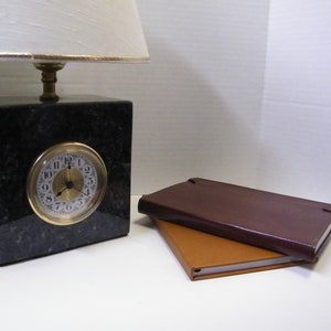 May include: A black granite clock with a white face and gold hands. The clock is on a table next to two brown leather-bound journals.