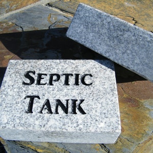 May include: A rectangular granite sign with the words "SEPTIC TANK" carved into the surface. The sign is light gray with dark gray speckles. A matching rectangular piece of granite is next to the sign.