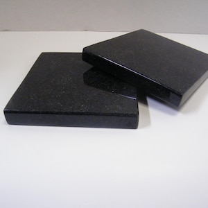 May include: Two black granite coasters with a polished finish. The coasters are square-shaped and have a slightly beveled edge. They are displayed on a white surface. These coasters are designed to protect surfaces from heat and moisture.
