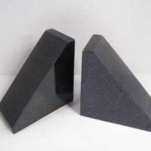 Puede incluir: Dos sujetalibros de granito negro. Cada sujetalibros tiene forma de prisma triangular con una parte superior plana y un acabado pulido. Los sujetalibros se exhiben sobre una superficie blanca.