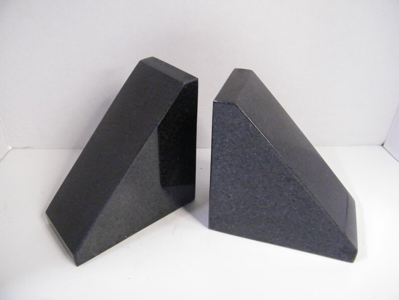Puede incluir: Dos sujetalibros de granito negro. Cada sujetalibros tiene forma de prisma triangular con una parte superior plana y un acabado pulido. Los sujetalibros se exhiben sobre una superficie blanca.