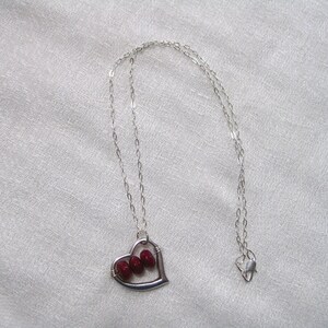 May include: A silver-toned necklace featuring a heart-shaped pendant. The pendant has a wire frame with three red beads inside. The chain is delicate and the clasp is visible. The necklace is displayed on a white, textured surface.