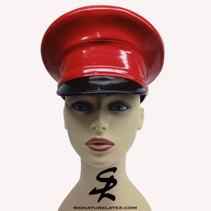Op de afbeelding: Een glanzende rode hoed met een zwarte rand, gepresenteerd op een mannequinhoofd. De hoed heeft een ronde kroon en een platte, cirkelvormige bovenkant. De mannequin heeft blauwe ogen en een tatoeage op de nek. De website signaturelatex.com staat onderaan afgedrukt.