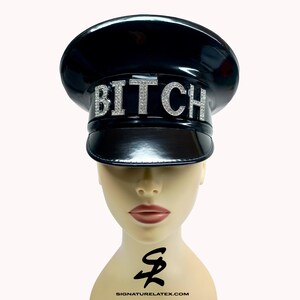 Op de afbeelding: Een zwarte, glanzende hoed met een ronde kroon en een klep. Het woord "BITCH" is in zilverkleurige, glinsterende letters over de voorkant geschreven. De hoed wordt getoond op een mannequinhoofd.