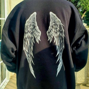 May include: Black hooded sweatshirt with a white angel wing design on the back. The wings are detailed with feather-like lines. The sweatshirt has a hood and long sleeves. The image is taken outdoors.