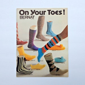 May include: A vintage advertisement titled "On Your Toes!" featuring various knitted socks and slippers. The socks showcase diverse patterns and colors, including argyle, stripes, and cable knit designs. The ad is from Bernat.