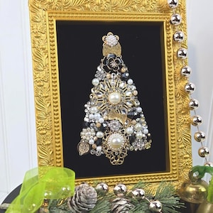 May include: A decorative wall art piece featuring a Christmas tree shape constructed from various vintage-style jewelry pieces, including pearls, rhinestones, and gold-tone metal accents, mounted inside an ornate, rectangular gold-colored frame.