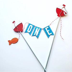 May include: A cake topper featuring two white sticks connected by a red and white string. Three blue pennant flags with white letters spell out the word ONE. Two red and white fishing bobbers are attached, with a small orange fish cutout.