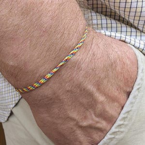 May include: A braided bracelet in a rainbow of colors, including red, yellow, green, and blue. The bracelet is worn on a wrist, with a neutral background.